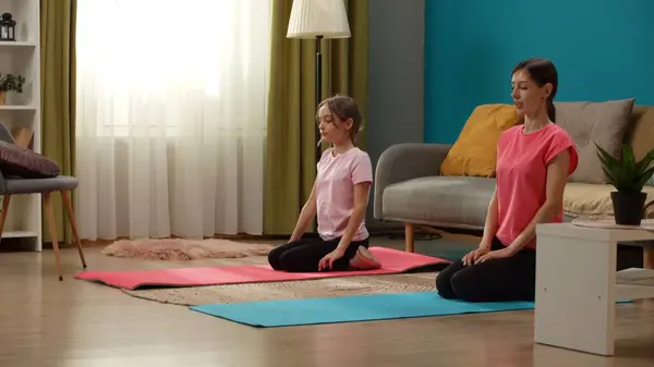 The footage shows a woman and a child sitting in a room on a fitness mat. They are demonstrating a yoga pose. Closing their eyes they meditate, concentrate, tune in. Medium shot.