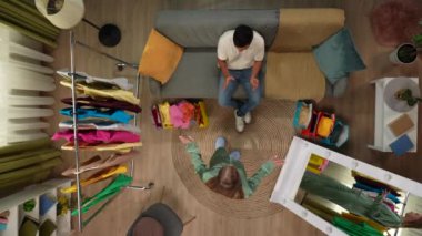 Alışveriş ve uygun yaratıcı reklam konsepti. Oturma odasının en üst manzarası. Kanepedeki adam, aynanın önünde yeni pijamalı kız, erkek kıyafetleri seviyor, kız kıyafetten memnun değil, tartışıyor..