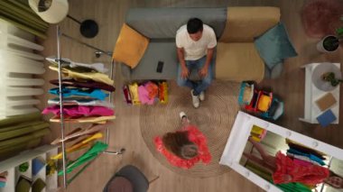 Alışveriş ve uygun yaratıcı reklam konsepti. Oturma odasının en üst manzarası. Adam kanepeye oturur, mutlu kız aynanın önünde yeni bir elbiseyle poz verir, adam kıyafeti sever ve başparmağını kaldırır..