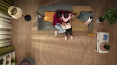 Bir anne ve çocuğun üstündeki karede bir odadaki kanepede oturuyorlar. Kızı bir kitap tutuyor ve kadın ona parmaklarıyla bir şey gösteriyor. Konuşuyorlar. Birlikte okuyorlar..