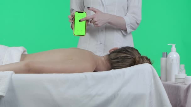医療広告コンセプト マッサージテーブルの女性の近くに クロマキーグリーンスクリーンでスマートフォンを保持するプロのセラピストが表示されます 広告エリア ワークスペースモックアップ — ストック動画
