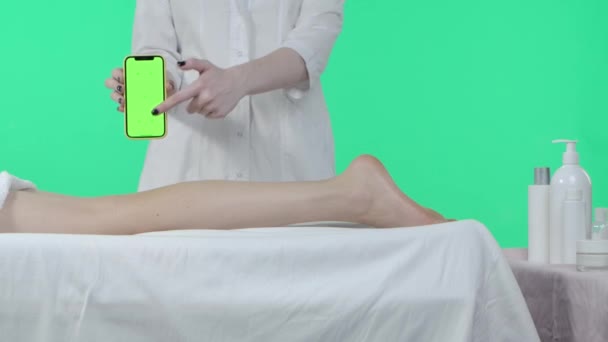 ボディトーナス広告コンセプト マッサージテーブルに女性の足を閉じ クロマキーグリーンスクリーンでスマートフォンを保持するプロのマッサージ師 スワイプディスプレイ 広告エリア ワークスペースモックアップ — ストック動画