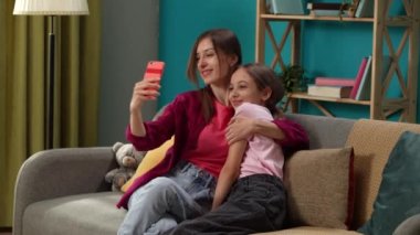 Yakın planda, bir kadın ve bir çocuk kanepede oturuyor, birbirlerine sarılıyorlar ve selfie çekiyorlar. Kameraya poz verip gülümsüyorlar. Sonra fotoğraflara bakıyorlar ve en iyisini seçiyorlar..