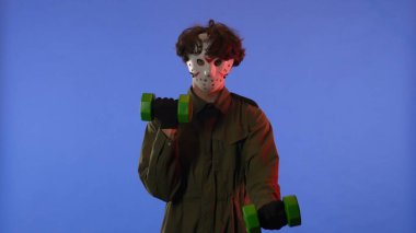 Beyaz maskeli bir adam kollarını mavi arka planda yeşil halterlerle sallıyor ve kırmızı ışık yanıyor. 13. Cuma 'dan manyak Jason Voorhees' in resmi. Cadılar Bayramı konsepti.
