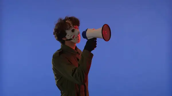 Man in white mask with loudspeaker in hands on blue background with red neon light. Image of maniac Jason Voorhees from Friday 13. Halloween celebration concept.