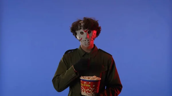 白いマスクの男は 赤いネオンライトで青い背景にポップコーンを食べています 金曜日の13からのマニアックジェイソン ヴァイヘイズのイメージ ハロウィーンのお祝いのコンセプト — ストック写真
