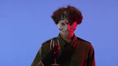 Beyaz maskeli bir adam kırmızı ışıklı, mavi arka planda kırmızı şaraplı bir bardak tutuyor. 13. Cuma 'dan manyak Jason Voorhees' in resmi. Yan görüş. Cadılar Bayramı konsepti