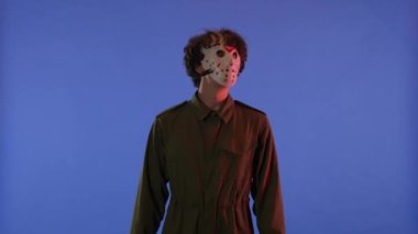 Beyaz maskeli adam etrafa bakıyor ve mavi arka planda kırmızı ışıklı parmaklarıyla iyi bir hareket sergiliyor. 13. Cuma 'daki Jason Voorhees manyağı. Cadılar Bayramı konsepti.