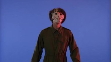 Beyaz maskeli bir adam etrafa bakıyor ve mavi arka planda kırmızı ışıklı parmakları olan jest yapıyor. 13. Cuma 'daki Jason Voorhees manyağı. Cadılar Bayramı konsepti