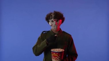 Beyaz maskeli bir adam mavi arka planda kırmızı ışıklı patlamış mısır yiyor. 13. Cuma 'dan manyak Jason Voorhees' in resmi. Cadılar Bayramı konsepti