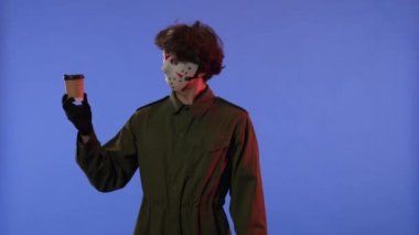 Beyaz maskeli ve bir fincan kahveli bir adam mavi arka planda kırmızı ışıklı bir jest gösteriyor. 13. Cuma 'daki Jason Voorhees manyağı. Cadılar Bayramı konsepti.