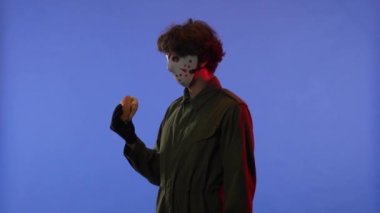 Beyaz maskeli bir adam hamburgeri kokluyor ve parmağını mavi arka planda kırmızı ışıkla işaret ediyor. 13. Cuma 'daki Jason Voorhees manyağı. Cadılar Bayramı konsepti.
