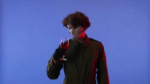 白いマスクの男は 赤いネオンライトで青い背景に赤いワインとグラスを保持しています 金曜日の13からのマニアックジェイソン ヴァイヘイズのイメージ サイドビュー ハロウィーンのお祝いのコンセプト — ストック動画