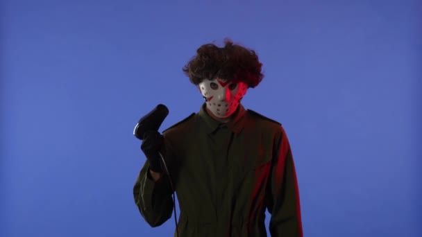 赤いネオン ライトが付いている青い背景のヘアドライヤーが付いている白いマスクの乾燥の毛の男 金曜日の13からのマニアックジェイソン ヴァイヘイズのイメージ ハロウィーンのお祝いのコンセプト — ストック動画