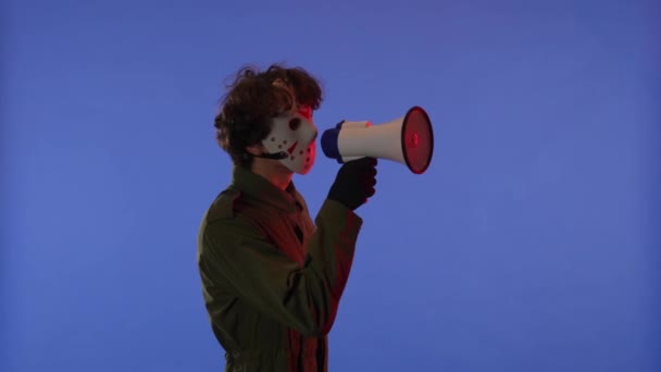 赤いネオン ライトが付いている青い背景のスピーカーが付いている白いマスクの男 金曜日の13からのマニアックジェイソン ヴァイヘイズのイメージ 彼は何かを言っている ハロウィーンのお祝いのコンセプト — ストック動画