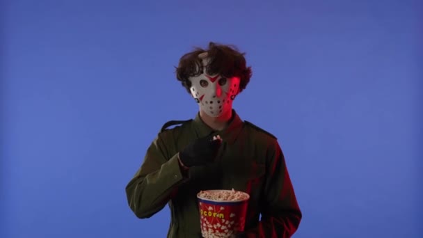 白いマスクの男は 赤いネオンライトで青い背景にポップコーンを食べています 金曜日の13からのマニアックジェイソン ヴァイヘイズのイメージ ハロウィーンのお祝いのコンセプト — ストック動画