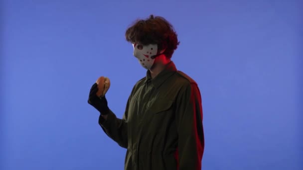 白いマスクの男がハンバーガーを嗅ぎ 赤いネオンライトで青い背景に向かって指を指差した 13日の金曜日からジェイソン ヴァイエス マニアックのイメージ ハロウィーンのお祝いのコンセプト — ストック動画