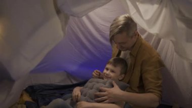 Oturma odasında kurulmuş bir çadırda babasının kollarında yatan bir oğul. Küçük bir çocuğu olan bir adam, çelenk ile aydınlatılmış bir çadırın içinde yastıklı bir şekilde oturmuş HDR BT2020 HLG materyaliyle