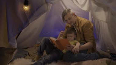 Aile zamanı konsepti. Oğluyla birlikte oturma odasındaki çadırda kitap okuyan bir baba. Oğlu sırt üstü babasının göğsüne yaslanmış yatıyordu ve babası kitabı işaret ettiğinde baş parmağını kaldırdı.