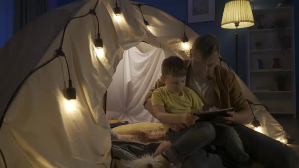 爸爸和儿子共享平板电脑 一起玩网络游戏 男人们竖起大拇指 儿子坐在父亲的膝上 靠近建在黑暗客厅里的花环帐篷 — 图库视频影像