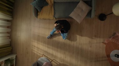 Oturma odasının zemininde, kanepenin yanında elinde akıllı bir telefonla oturan bir adamın en üst görüntüsü. Telefonda konuşan adam.