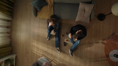 Oturma odasının zemininde bir şişe alkol, bardak ve küllükle oturan bir çiftin en üst görüntüsü. Alkol yardımıyla sorunlarından kaçan bir çift.