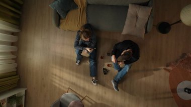 Oturma odasının zemininde bir şişe alkol, bardak ve küllükle oturan bir çiftin en üst görüntüsü. Bir kadın elinde sigara tutuyor. Sorunlarından kaçan bir çift.