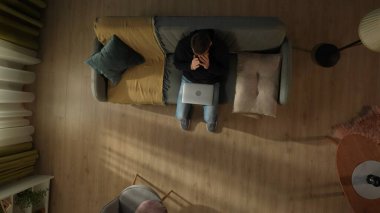 Oturma odasındaki kanepede oturan yorgun bir adamın dizüstü bilgisayarı kapalıyken en iyi görüntüsü. Adam gözlerini ovuşturuyor. Yorgunluk, acı, gözlerde yarıklar.