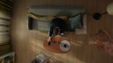 Oturma odasındaki kanepede oturan bir adamın bir şişe alkol ve bardakla kahve masasının önünde görüntüsü. Adam gergin bir şekilde telefonuna bakıyor, bir telefon ya da mesaj bekliyor.