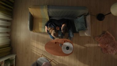 Oturma odasındaki kanepede oturan bir çiftin bir şişe alkol ve bardakla birlikte kahve masasının önündeki görüntüsü. Erkek sarılır ve perişan kadını teselli eder.