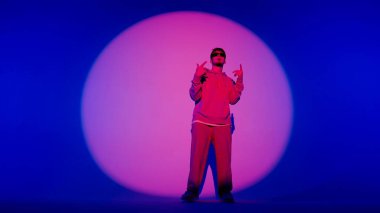 Genç bir adam, üzerinde pembe bir ışıkla mavi bir arka planda duruyor. Daire çiziyor. Dans hareketini gösteriyor. Sokak tarzı ve gözlüklü kıyafetler giymiş..