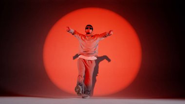 Kırmızı arkaplanda genç bir adam duruyor. Spot ışığı parlıyor. Daire çiziyor. Dans hareketini, atlamayı gösteriyor. Sokak tarzı giyinmiş ve gözlüklü..