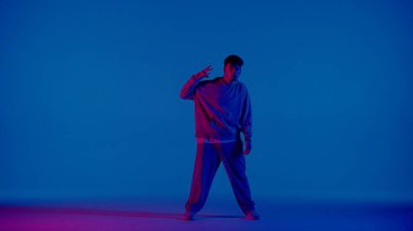 Genç bir adam mavi arka planda duruyor ve kameraya bakıyor. Yandan ona pembe bir ışık yansıyor ve bir tarafı aydınlatıyor. Dans eden ve dans eden bir dansçı..