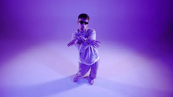 There is a man standing on a purple background. The camera is slightly overhead. Demonstrates a dance movement, rays of light shine on him. Dressed in street style clothing. He is plastic, rhythmic.