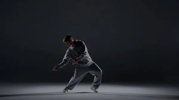 A young man stands against a dark background. He is highlighted by a white light from below. Creates a light haze. Demonstrates a dance movement by leaning to the side. He is plastic, rhythmic.