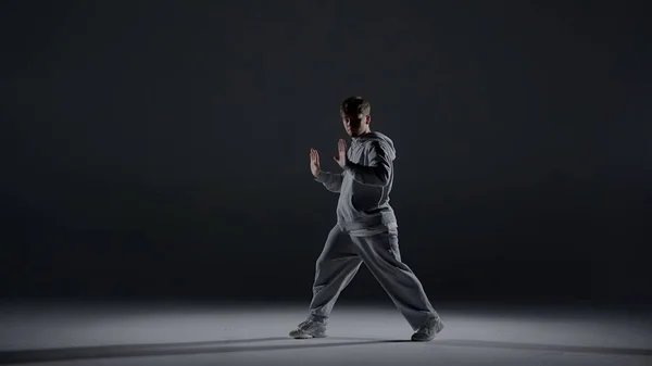 A young man stands against a dark background. He is highlighted by a white light from below. Creates a light haze. Demonstrates a dance movement by extending his arms forward. He is plastic, rhythmic.