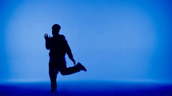 青と白の背景のフレームには 若い男がシルエットに立っています ダンスの動きをデモンストレーションし 片足を上げる 彼はプラスチックでリズム的だ 彼はストリートスタイルの服を着ている メディア — ストック写真