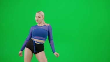 Yeşil arka planda çerçevelenmiş, kromakey. Genç, çekici bir kadın ayakta duruyor, parlak, seksi kıyafetler giyiyor ve ellerini kullanarak bir dans hareketi sergiliyor. Dikkatlice kameraya bakıyor..