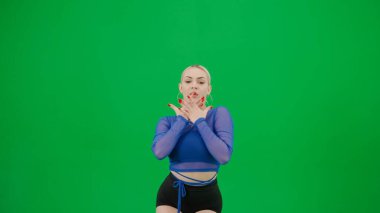 Yeşil arka planda çerçevelenmiş, kromakey. Genç, çekici bir kadın ayakta duruyor, parlak, seksi kıyafetler giyiyor ve ellerini kullanarak bir dans hareketi sergiliyor. Dikkatlice kameraya bakıyor..