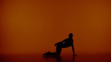 Kırmızı, turuncu arka planda çerçevelenmiş. Silüet giymiş, açık giysiler giymiş bir kadın. Dans hareketini gösteriyor, poz veriyor. Diz çöküp elini yere koydu. Seksi, plastik..