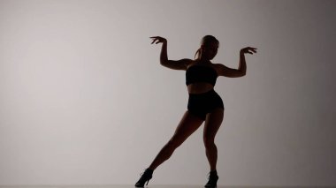 Çerçeve gri bir arkaplanda. Kadın biraz karamsar. Açık kıyafetler ve yüksek topuklu ayakkabılar giyiyor. Dans hareketini, duruşu, bacakları ve kolları ayırmayı gösteriyor. Seksi, plastik..