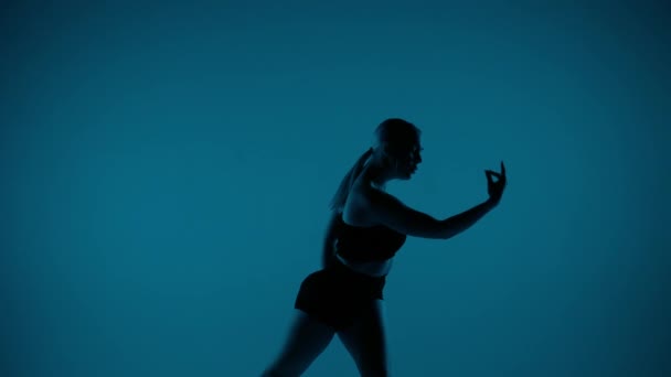 青い背景に近いショットで オープンな服を着た少女が立ってカメラを見ている 手と足でダンスの動きを示しています 彼女はセクシーで プラスチック — ストック動画
