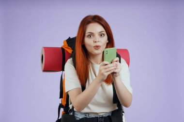 Turizm ve aktif boş zaman reklam konsepti. Kadın gezginin portresi. Sırt çantalı kadın turist akıllı telefondan fotoğraf çekiyor, şaşkın surat. Mor arkaplanda izole.