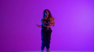 Modern koreografi ve dans stili yaratıcı konsept. Kadın dansçı portresi. Siyah pantolonlu ve ceketli çekici kadın caz funk koreografisi yapıyor, mor arka planda izole edilmiş neon ışıklı..