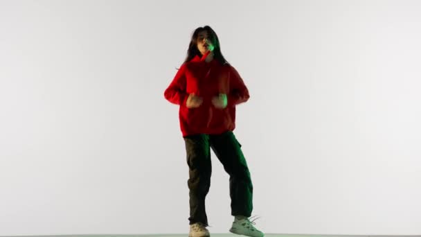 現代の振付とダンススタイルのクリエイティブコンセプト 女性ダンサーの肖像画 白い背景緑のネオンライトで隔離されたジャズファンクの振付を踊る赤いフーディーの魅力的な女性 — ストック動画