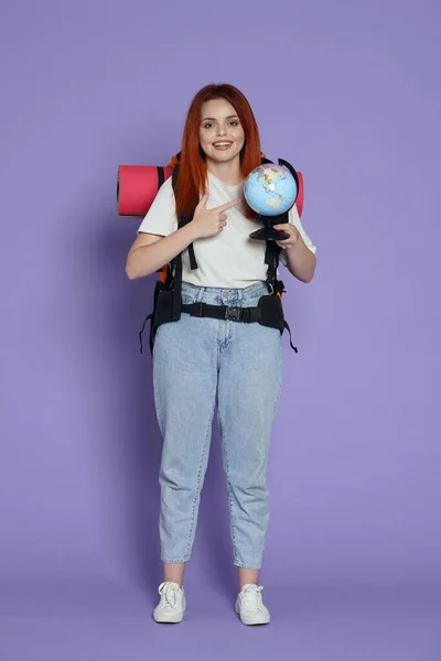 旅游和积极的业余时间广告概念 女旅行家的肖像穿着休闲装 背着背包的女游客微笑着 举着指向地图的地球仪 被紫色背景隔离 — 图库照片