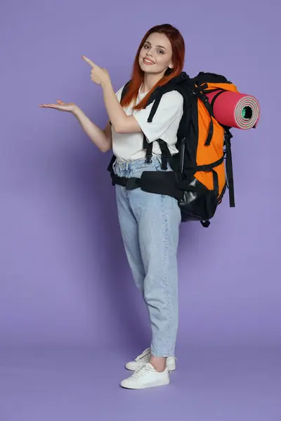観光とアクティブなフリータイム広告コンセプト 女の子旅行者の肖像画 バックパックを指すカジュアルな女性観光客 広告のためのスペース 紫色の背景に隔離される — ストック写真