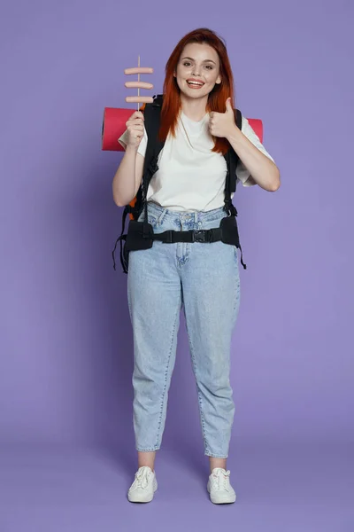旅游和积极的业余时间广告概念 女旅行家的肖像女游客穿着休闲装 背包里拿着腊肠棒 竖起大拇指 被紫色背景隔离 — 图库照片