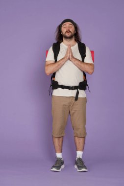 Turizm ve aktif boş zaman reklam konsepti. Erkek gezginin portresi. Sırt çantalı, sıradan giyinmiş, gözleri kapalı dua eden bir turist. Mor arkaplanda izole.