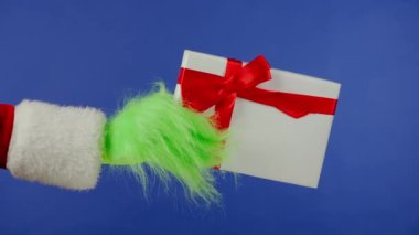 Grinchs 'in yeşil saçlı eli, izole edilmiş mavi arka planda kırmızı fiyonklu beyaz bir hediye kutusu tutuyor. Hediye kaçıran kostüm. Noel ve Yeni Yıl kutlaması konsepti. Mavi ekran, krom anahtar.